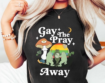 Könnte beinhalten: Schwarzes T-Shirt mit einem weißen Motiv von drei Fröschen, die um einen Pilz mit einem Regenbogen tanzen, und dem Text "Gay The Pray Away".