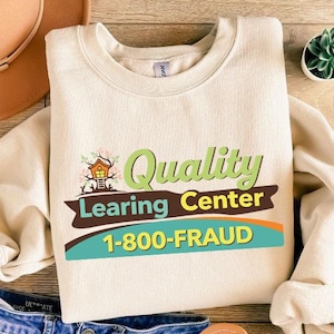 May include: Cream-colored sweatshirt with the text "Quality Learning Center 1-800-FRAUD" in green, brown, and teal. The sweatshirt is laid flat with a pair of blue jeans, a brown hat, and sandals.