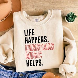 May include: Cream-colored sweatshirt with the text "LIFE HAPPENS. CHRISTMAS MUSIC HELPS." in black and red. The sweatshirt is paired with blue denim shorts, a brown hat, sandals, and a small succulent plant.
