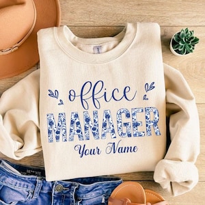 May include: Cream-colored sweatshirt with the words "office MANAGER Your Name" in blue script and floral print. The sweatshirt is paired with blue jeans, a brown hat, and sandals. A small succulent plant is also visible.