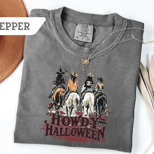 May include: A grey Comfort Colors t-shirt with a graphic of four riders on horseback and the text "Howdy Halloween" in red. A brown hat and a silver necklace are also visible.