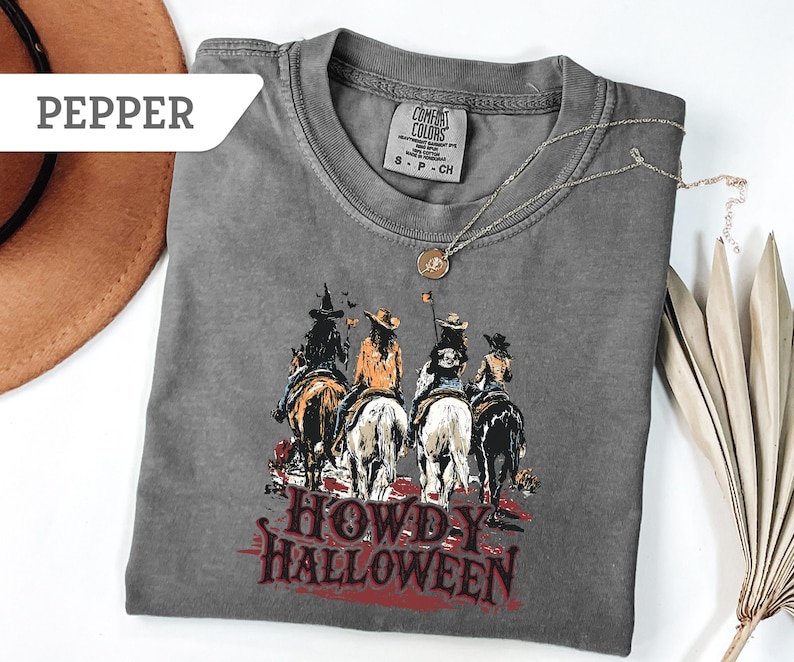 May include: A grey Comfort Colors t-shirt with a graphic of four riders on horseback and the text "Howdy Halloween" in red. A brown hat and a silver necklace are also visible.