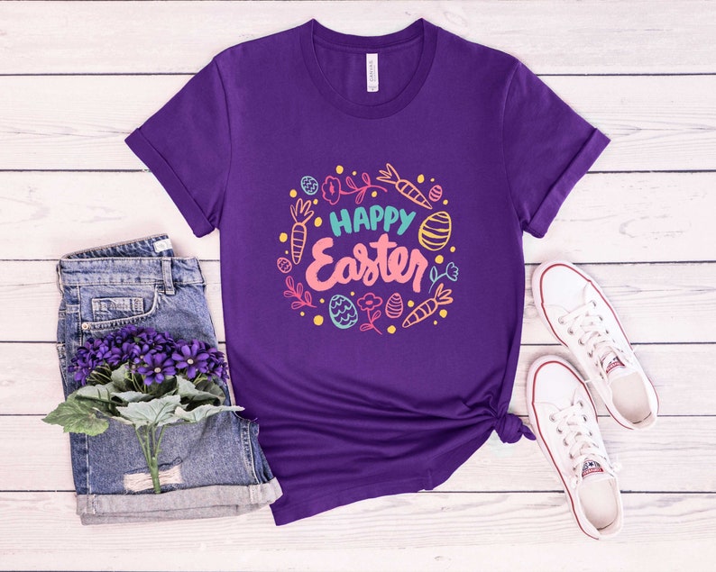 May include: Purple t-shirt with a white crew neck and a colorful graphic design that says "Happy Easter" surrounded by carrots, eggs, and flowers.