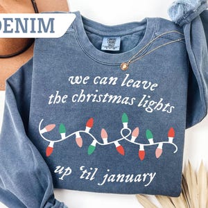 May include: Denim blue sweatshirt with the text "we can leave the christmas lights up til january" and a string of Christmas lights in red, green, and pink. A gold necklace is draped over the sweatshirt.