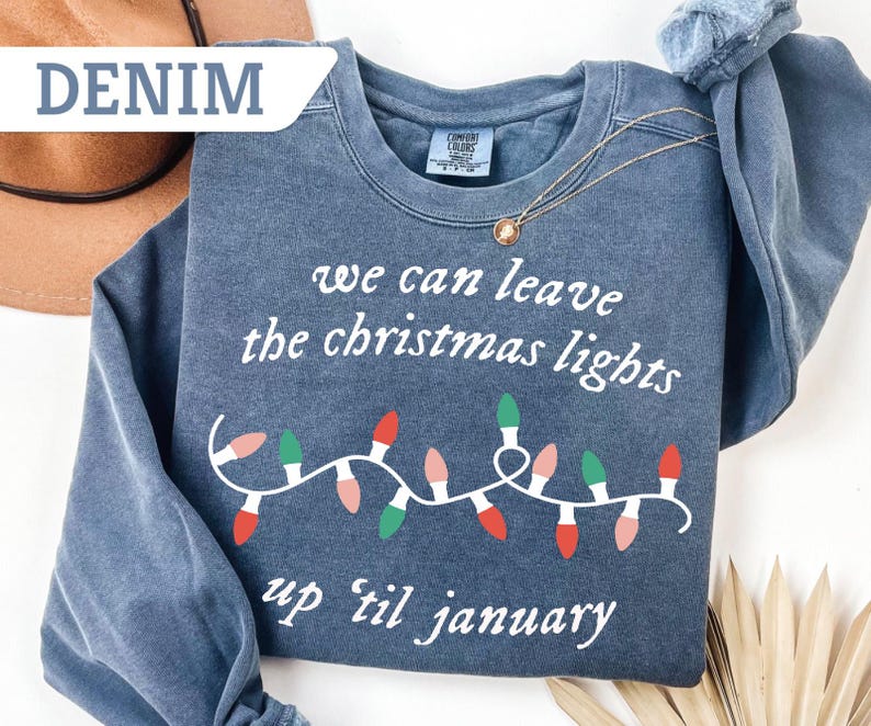 May include: Denim blue sweatshirt with the text "we can leave the christmas lights up til january" and a string of Christmas lights in red, green, and pink. A gold necklace is draped over the sweatshirt.