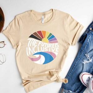 May include: A beige t-shirt with a graphic design of a rainbow sun and waves with the text "The Future is Inclusive".