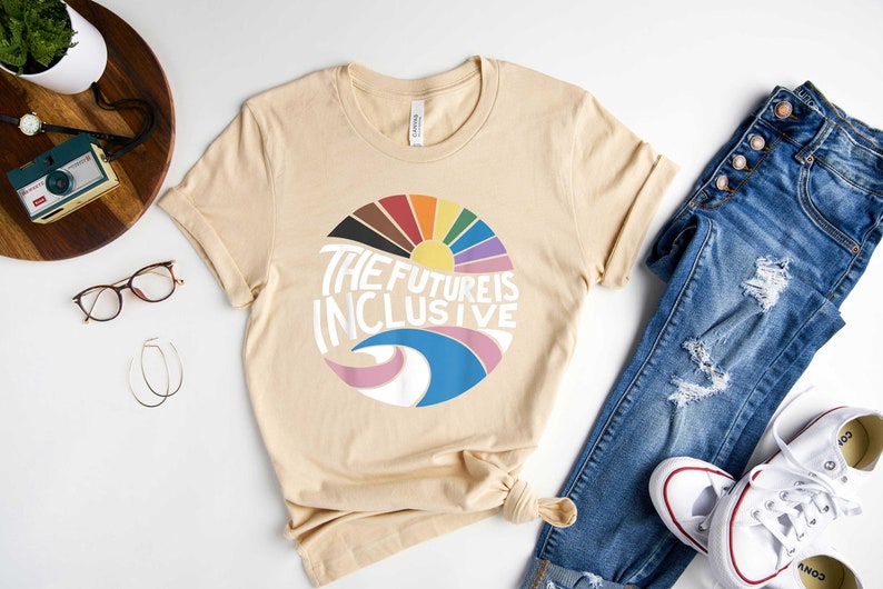 May include: A beige t-shirt with a graphic design of a rainbow sun and waves with the text "The Future is Inclusive".