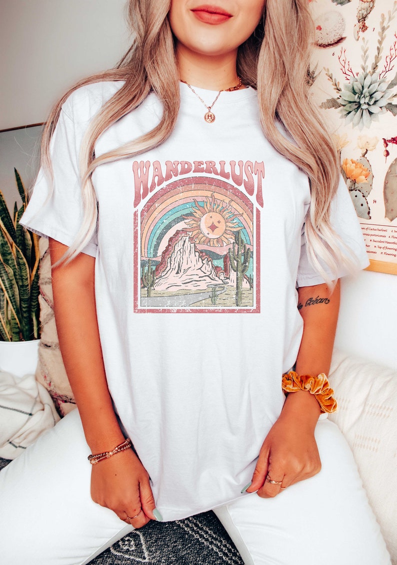 May include: White t-shirt with a vintage-style graphic of a mountain range, sun, and rainbow. The text "WANDERLUST" is printed in pink above the graphic.