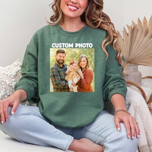 Puede incluir: Sudadera verde con una foto familiar impresa en la parte delantera. La foto muestra a una familia de tres. El texto "CUSTOM PHOTO" est&aacute; impreso encima de la foto en un rect&aacute;ngulo blanco. La persona de la imagen lleva vaqueros azul claro.