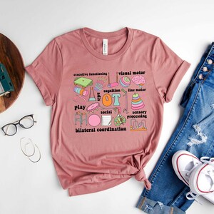 May include: A pink t-shirt with a graphic design of various colorful illustrations and text related to occupational therapy. The text includes "OT", "play", "cognition", "sensory processing", "executive functioning", "visual motor", "fine motor", "social", and "bilateral coordination".