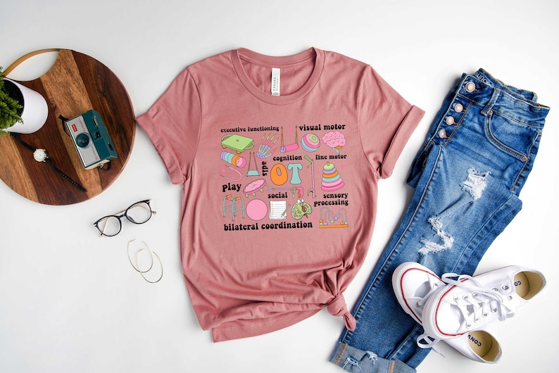 May include: A pink t-shirt with a graphic design of various colorful illustrations and text related to occupational therapy. The text includes "OT", "play", "cognition", "sensory processing", "executive functioning", "visual motor", "fine motor", "social", and "bilateral coordination".