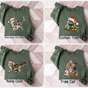 May include: Olive green sweatshirts with cat-themed Christmas designs. Each sweatshirt features a different cat breed wearing a Santa hat and Christmas lights. The breeds include American Shorthair, Scottish Fold, Maine Coon, and a Tree Cat design.