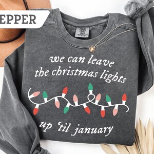 May include: A dark gray sweatshirt with the text "we can leave the christmas lights up 'til january" in white. A string of Christmas lights in red, green, and pink is below the text. A gold necklace is draped on the sweatshirt.