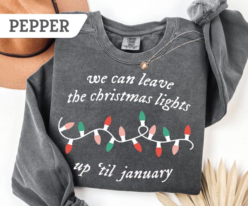 May include: A dark gray sweatshirt with the text "we can leave the christmas lights up 'til january" in white. A string of Christmas lights in red, green, and pink is below the text. A gold necklace is draped on the sweatshirt.