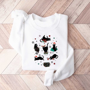 May include: White sweatshirt featuring a festive design of black cats celebrating Christmas. The cats are wearing Santa hats and are surrounded by stars, snowflakes, and Christmas lights.