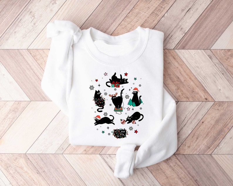 May include: White sweatshirt featuring a festive design of black cats celebrating Christmas. The cats are wearing Santa hats and are surrounded by stars, snowflakes, and Christmas lights.