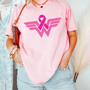 May include: Pink t-shirt with a pink ribbon and a Wonder Woman logo. The logo is a stylized 'W' with wings and a pink ribbon in the center.