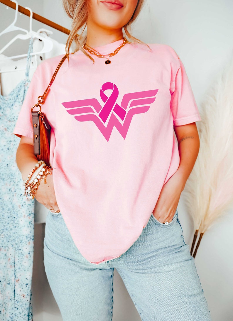 May include: Pink t-shirt with a pink ribbon and a Wonder Woman logo. The logo is a stylized 'W' with wings and a pink ribbon in the center.