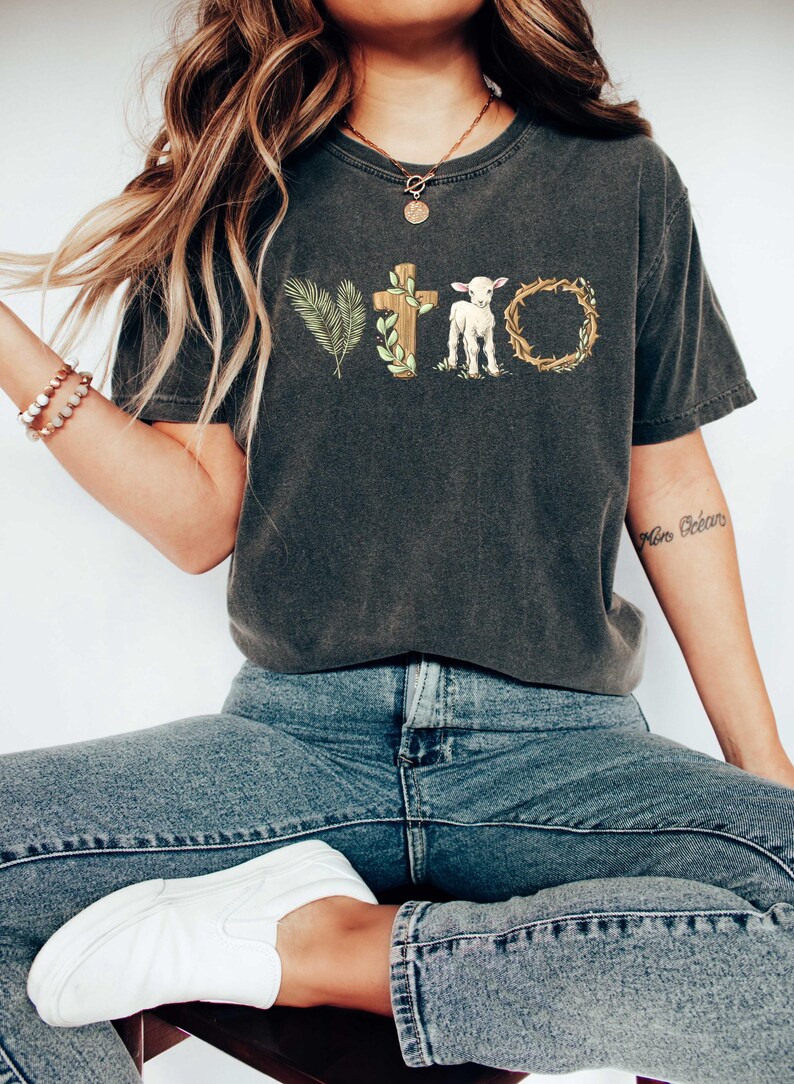 May include: A dark gray t-shirt with a graphic design of a cross, a lamb, and a crown of thorns. The graphic is in shades of brown, green, and white.