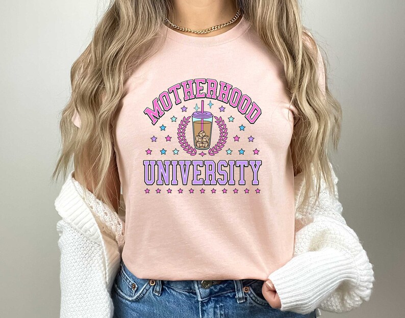 May include: Pink t-shirt with the text "MOTHERHOOD UNIVERSITY" in pink and purple letters. A cartoon image of a boba tea cup with brown liquid, ice cubes, and a straw is in the center of the text. The text is surrounded by pink and blue stars.