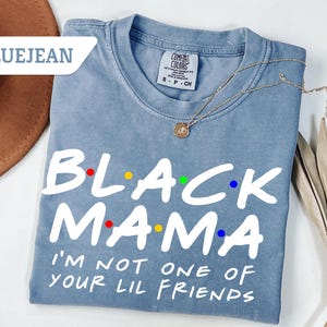 May include: Bluejean Comfort Colors t-shirt with the text "BLACK MAMA" in white, with colourful dots. Below the text is "I'M NOT ONE OF YOUR LIL FRIENDS" in white. A gold necklace is draped across the shirt.