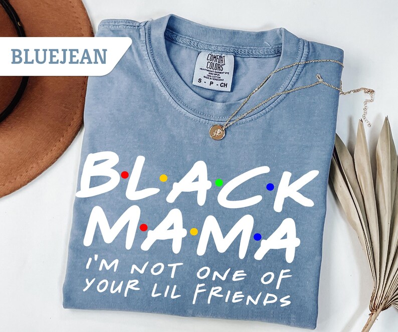 May include: Bluejean Comfort Colors t-shirt with the text "BLACK MAMA" in white, with colourful dots. Below the text is "I'M NOT ONE OF YOUR LIL FRIENDS" in white. A gold necklace is draped across the shirt.