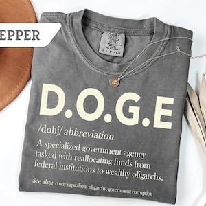May include: A gray t-shirt with the word "D.O.G.E" in large, off-white letters. Below the word is a definition. A silver necklace with a pendant rests on the shirt. A brown hat is visible in the background.