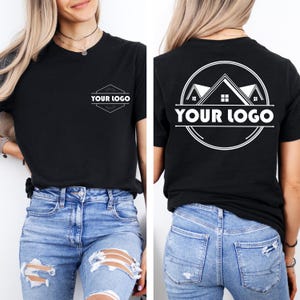 May include: A black t-shirt featuring a white graphic design. The front displays a small hexagonal logo with the text "YOUR LOGO". The back features a large circular graphic with a house roof illustration and the text "YOUR LOGO" in bold letters.