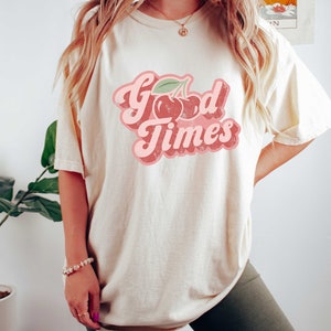 Pu&ograve; includere: T-shirt color crema con la scritta "Good Times" in un font retr&ograve;, con una grafica di ciliegie. La maglietta ha una vestibilit&agrave; ampia e il design &egrave; nei toni del rosa e del rosso.