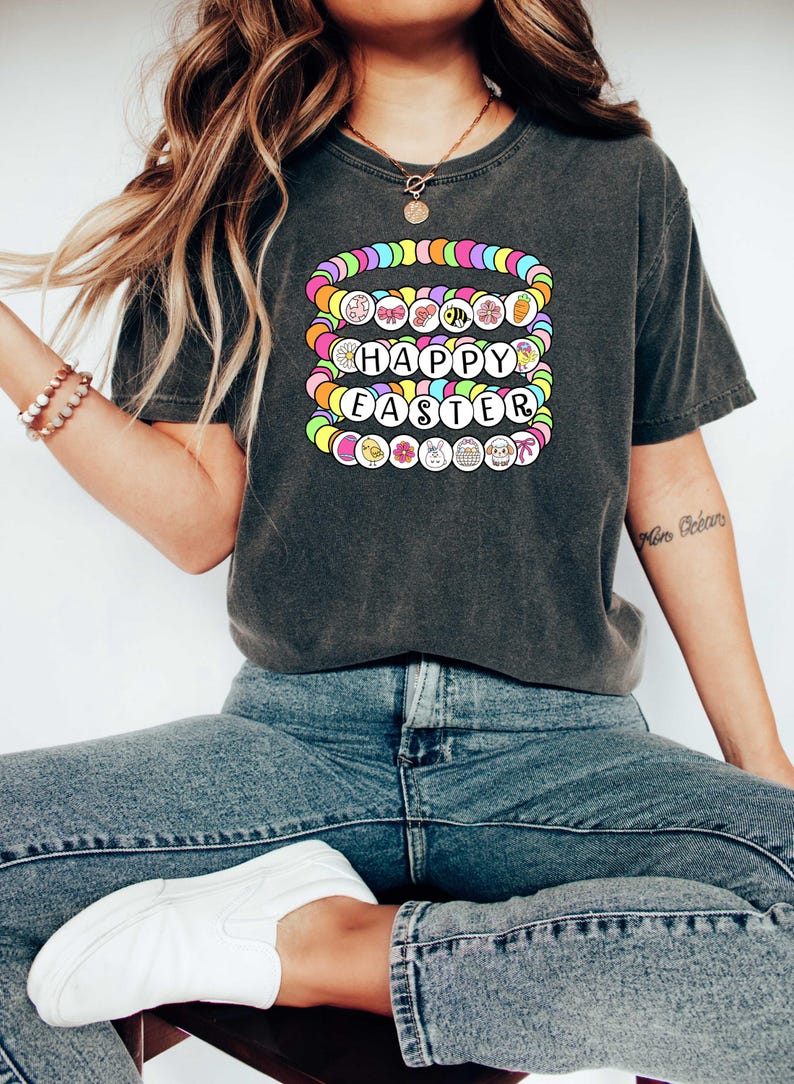 May include: A dark grey t-shirt with a graphic of colorful beads spelling out "Happy Easter". The beads are decorated with Easter-themed icons like bunnies, eggs, and flowers.
