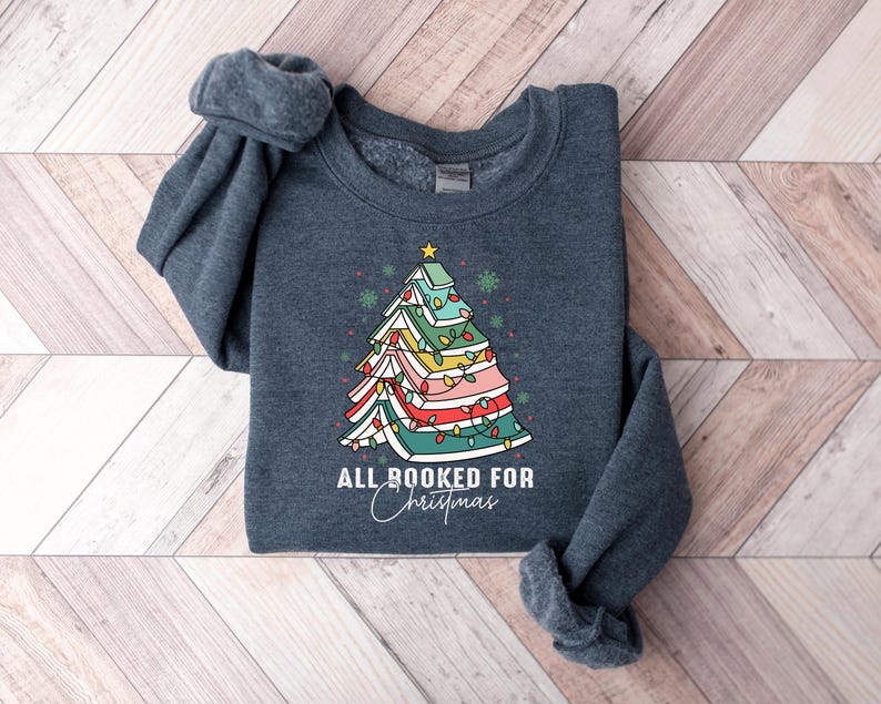 May include: A dark gray sweatshirt featuring a colorful Christmas tree design made of books, with string lights and a star on top. The text "ALL BOOKED FOR Christmas" is written below the tree in a script font.