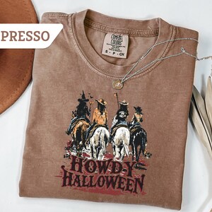 May include: Espresso-coloured t-shirt featuring a graphic of four figures on horseback, with the text "HOWDY HALLOWEEN" in red. The shirt has a crew neck and short sleeves. A silver necklace with a pendant is draped on the shirt.