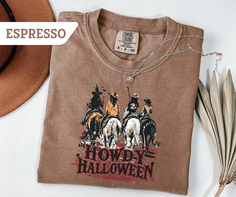 May include: Espresso-coloured t-shirt featuring a graphic of four figures on horseback, with the text "HOWDY HALLOWEEN" in red. The shirt has a crew neck and short sleeves. A silver necklace with a pendant is draped on the shirt.