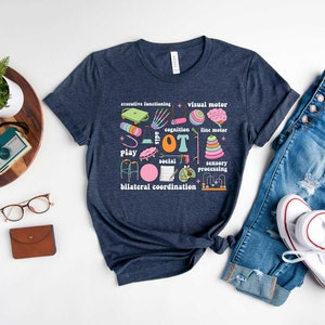 May include: A navy blue t-shirt with a graphic design of occupational therapy tools and the text "OT" in pink. The graphic includes a walker, a ball, a drum, a brain, a hand, a pencil, and other tools.