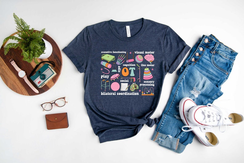 May include: A navy blue t-shirt with a graphic design of occupational therapy tools and the text "OT" in pink. The graphic includes a walker, a ball, a drum, a brain, a hand, a pencil, and other tools.