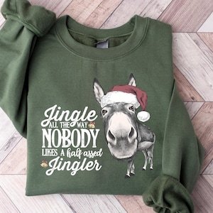 May include: Green sweatshirt featuring a donkey wearing a Santa hat. The text reads "Jingle All The Way Nobody Likes A Half Assed Jingler" in white script. The sweatshirt has long sleeves and a crew neck, perfect for the holiday season.