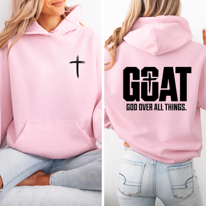 Pode incluir: Um moletom rosa claro com um bolso frontal e capuz. A frente apresenta uma cruz preta. A parte de tr&aacute;s do moletom exibe a palavra "GOAT" com uma cruz no "O" e o texto "GOD OVER ALL THINGS."