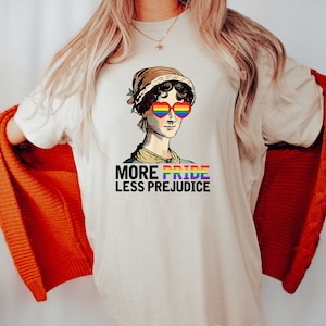 May include: A white t-shirt featuring a graphic of a woman wearing rainbow heart-shaped sunglasses. The text "More Pride Less Prejudice" is printed below the graphic.