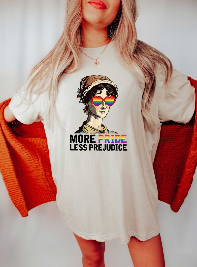 May include: A white t-shirt featuring a graphic of a woman wearing rainbow heart-shaped sunglasses. The text "More Pride Less Prejudice" is printed below the graphic.