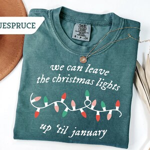 May include: A teal Comfort Colors t-shirt with the text "we can leave the christmas lights up 'til january" and a string of Christmas lights. A gold necklace with a pendant is on the shirt.