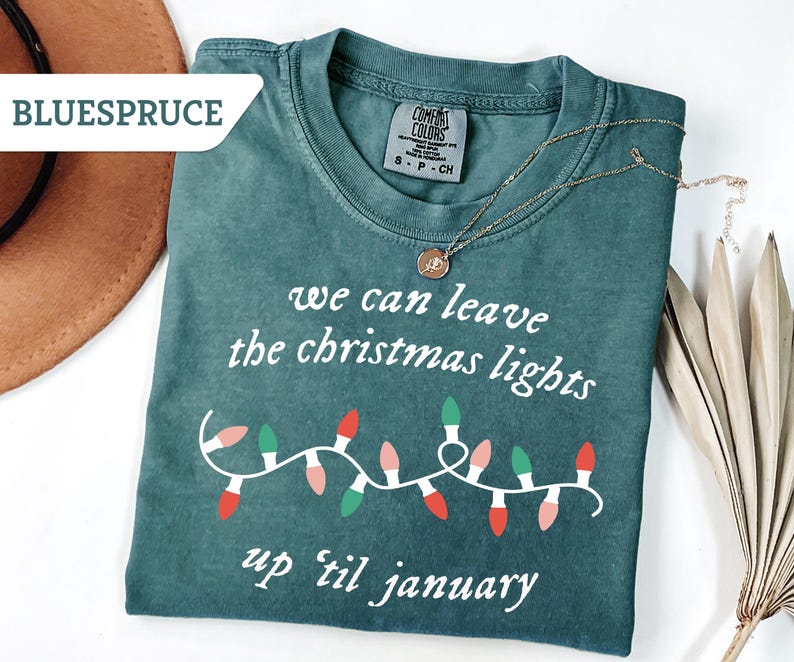May include: A teal Comfort Colors t-shirt with the text "we can leave the christmas lights up 'til january" and a string of Christmas lights. A gold necklace with a pendant is on the shirt.