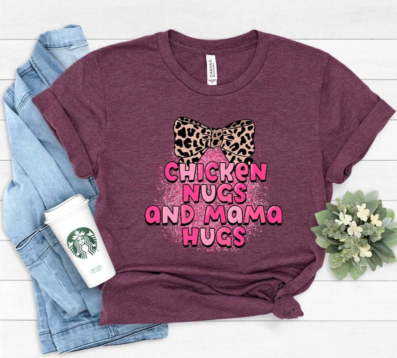 May include: A maroon t-shirt with a pink glitter design that says "Chicken Nugs and Mama Hugs". A leopard print bow is above the text.