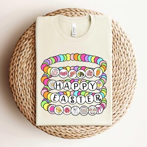 May include: A light beige t-shirt with a colorful graphic of a stack of bracelets that spell out "Happy Easter". The bracelets are decorated with various Easter-themed charms, including flowers, bunnies, eggs, and chicks.