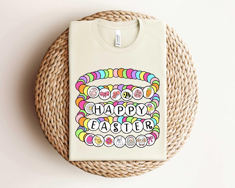 May include: A light beige t-shirt with a colorful graphic of a stack of bracelets that spell out "Happy Easter". The bracelets are decorated with various Easter-themed charms, including flowers, bunnies, eggs, and chicks.