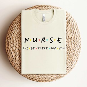 Puede incluir: Una camiseta beige con el texto "NURSE" en letras negras con corazones coloridos entre las letras. El texto "I'll be there for you" est&aacute; impreso en negro debajo.
