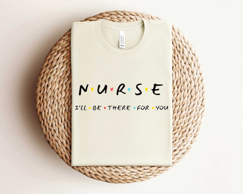 Puede incluir: Una camiseta beige con el texto "NURSE" en letras negras con corazones coloridos entre las letras. El texto "I'll be there for you" est&aacute; impreso en negro debajo.
