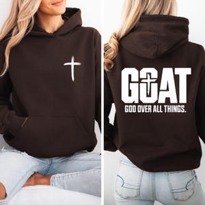 Pode incluir: Um moletom marrom escuro com uma cruz branca na frente e a palavra "GOAT" com uma cruz no "O" nas costas. O texto "GOD OVER ALL THINGS" est&aacute; abaixo da palavra "GOAT".