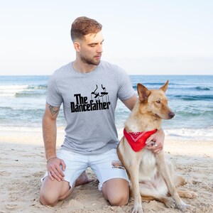 Pode incluir: Um homem vestindo uma camiseta cinza com o texto "The Dancefather" e uma silhueta preta de uma pessoa dan&ccedil;ando. Ele est&aacute; ajoelhado em uma praia de areia com um cachorro marrom usando uma bandana vermelha.
