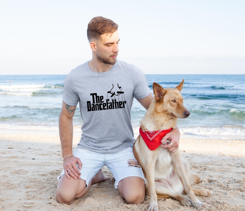 Pode incluir: Um homem vestindo uma camiseta cinza com o texto "The Dancefather" e uma silhueta preta de uma pessoa dan&ccedil;ando. Ele est&aacute; ajoelhado em uma praia de areia com um cachorro marrom usando uma bandana vermelha.