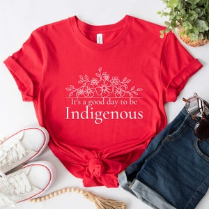 May include: Red t-shirt with a white floral design and the text "It's a good day to be Indigenous".